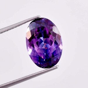 Puede incluir: Una piedra preciosa ovalada de color púrpura con un corte facetado, sostenida por unas pinzas sobre un fondo blanco.