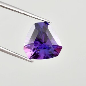 Peut inclure: Une pierre précieuse triangulaire facettée, tenue par une pince. La gemme présente un dégradé de couleurs, passant du violet profond à des nuances plus claires de violet et de rose. La taille crée de multiples facettes.