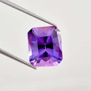 May include: A rectangular-cut gemstone, held by tweezers. The gem displays a gradient of colour, from light pink to deep purple. The facets of the gem reflect light. A beautiful example of a precious stone.
