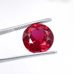 May include: A deep red gemstone with a round cut and many facets. The gemstone is held in place by a pair of tweezers.