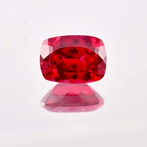 May include: A deep red gemstone with a cushion cut. The gemstone is reflecting light and has a shiny finish.