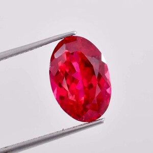 May include: A vibrant red oval-shaped gemstone held by tweezers. The gemstone is faceted and has a high polish.