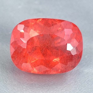 Puede incluir: Una gema roja brillante, facetada, con una forma ligeramente ovalada. La gema tiene una superficie lisa y pulida y está colocada sobre un fondo gris.