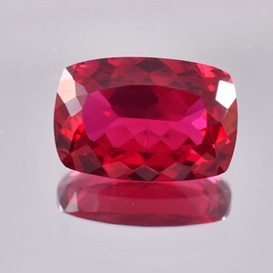 Puede incluir: Una piedra preciosa de color rojo intenso con un corte cojín y un alto pulido. La piedra preciosa se coloca sobre un fondo blanco.