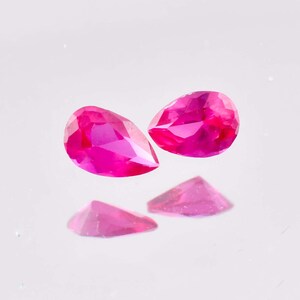 Peut inclure: Deux pierres précieuses roses vif en forme de poire, avec de multiples facettes, sont présentées sur un fond blanc. Deux plus petits reflets triangulaires des pierres sont visibles en dessous. Les pierres sont probablement des rubis ou des pierres précieuses similaires.