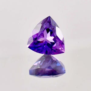 Puede incluir: Una piedra preciosa de amatista tallada en forma triangular con una faceta en forma de estrella en el centro. La piedra preciosa es de color morado oscuro y tiene un alto brillo.