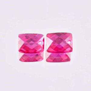 May include: Two faceted, square-shaped, hot pink beads with a hole drilled through each. The beads are highly reflective, with a mirrored surface beneath them. The beads are likely for jewelry making.