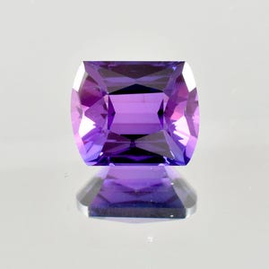 May include: A rectangular-cut gemstone in shades of purple. The gem's facets reflect light, creating a sparkling effect. The gem is set against a light background. The gem's dimensions are approximately 1.5 cm x 1 cm.