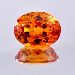 May include: A bright orange oval-shaped gemstone with a faceted cut. The gemstone is reflecting light and has a shiny finish.