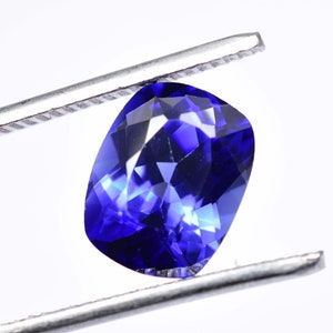 May include: A cushion-cut, deep blue gemstone is held by silver tweezers against a white backdrop. The gem's facets reflect light, creating a brilliant display of colour and sparkle. The gem is a vibrant shade of blue, similar to sapphire.