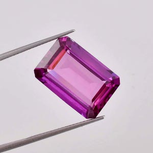 May include: A vibrant purple gemstone with a rectangular emerald cut. The gemstone is held in a pair of tweezers.