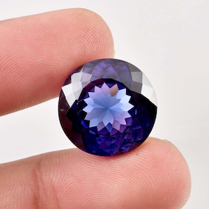 Puede incluir: Una gema redonda y facetada, de color púrpura intenso, con un diseño de estrella en el centro. La gema se sostiene entre dos dedos, mostrando su superficie pulida y su intrincado diseño. Las facetas reflejan la luz, creando un efecto brillante.