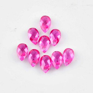 May include: A collection of ten faceted, teardrop-shaped, hot pink acrylic beads. Each bead has a hole at the top for stringing. The beads are arranged on a white surface, showcasing their vibrant color and cut.