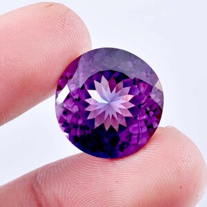 Puede incluir: Una piedra preciosa de amatista redonda y facetada con un patrón de estrella en el centro. La piedra preciosa es de color morado oscuro.