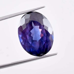 Könnte beinhalten: Ein ovaler Edelstein in tiefem Lila mit facettiertem Schliff. Der Edelstein wird von einer Pinzette vor einem weißen Hintergrund gehalten.