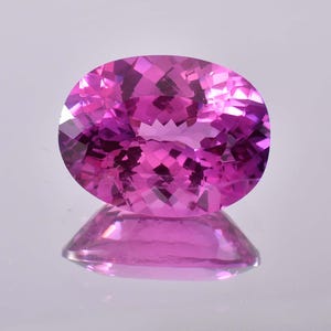 May include: A vibrant pink oval-shaped gemstone with a faceted cut. The gemstone is reflecting light and has a high level of brilliance.