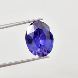 May include: A deep blue, oval-cut gemstone, held by silver tweezers. The faceted surface of the gem reflects light, displaying varying shades of blue. The background is plain white, accentuating the gemstone's colour and form.