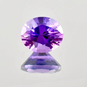 May include: A round-cut gemstone with a deep purple colour. The facets of the gem reflect light, creating a sparkling effect. The gemstone is set against a light background, with its reflection visible below.