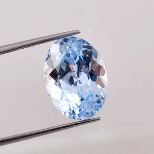 Puede incluir: Una gema ovalada de color azul claro sujeta con pinzas. La gema tiene muchas facetas, que reflejan la luz. El fondo es de color gris claro liso.