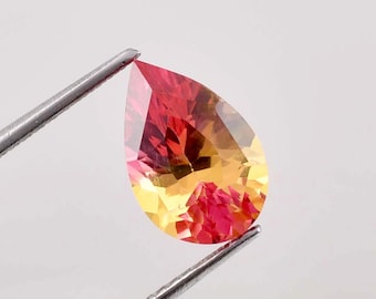 Può includere: Una pietra preziosa a forma di pera con un gradiente di colori rosso e giallo. La pietra preziosa è tenuta da una pinzetta.