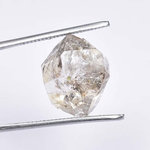 Puede incluir: Un cristal de diamante octaédrico transparente y facetado con inclusiones, sostenido por unas pinzas sobre un fondo blanco.