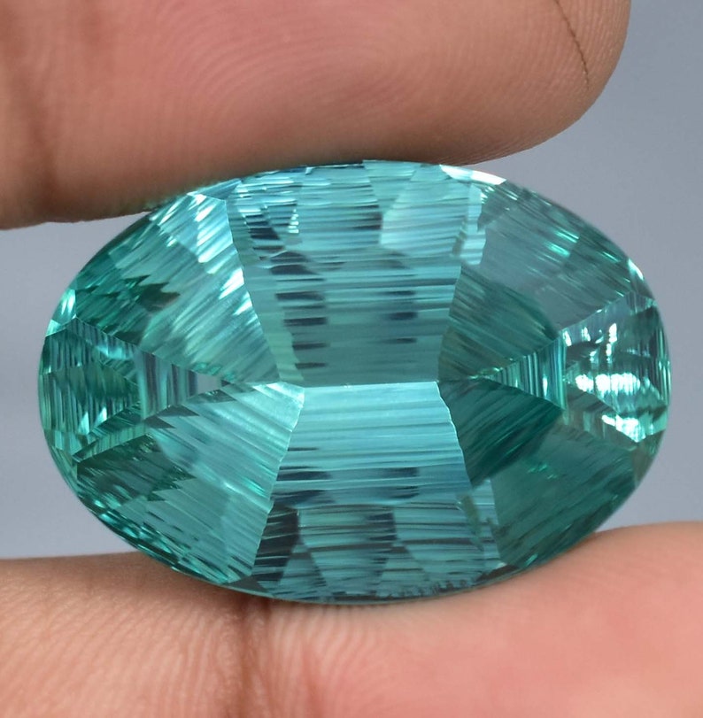 May include: A large, oval-shaped, faceted gemstone with a teal blue color. The gemstone has a striated pattern and is cut in a unique style.