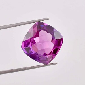 Puede incluir: Una piedra preciosa de talla cojín con un tono morado intenso. La gema está sujeta por unas pinzas sobre un fondo blanco.