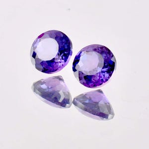 Puede incluir: Dos piedras preciosas de amatista redondas y facetadas con un degradado de color púrpura y púrpura claro. Las piedras preciosas están sobre una superficie blanca.