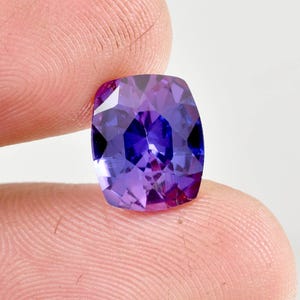 May include: A faceted, cushion-cut gemstone with a vibrant colour gradient. The gem transitions from deep violet to a lighter purple hue, with flashes of blue. The stone is held between fingers, showcasing its polished facets.