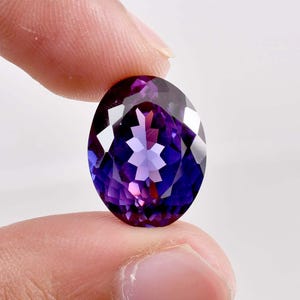 以下が含まれることがあります： 深い紫色の色合いから青とピンクに変化する、楕円形の宝石。指でつまんでおり、そのファセットと輝きを際立たせています。宝石のカットは、内部に星のような模様を明らかにしています。