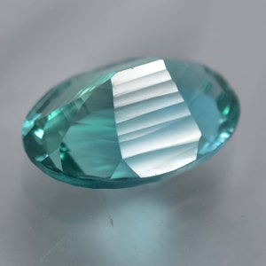 May include: A teal blue oval-shaped gemstone with a faceted cut. The gemstone is reflecting light, showing its facets.