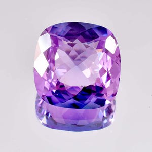 Puede incluir: Una gema facetada, de talla cojín, con tonos púrpuras. Las facetas de la gema reflejan la luz, creando un efecto brillante. La piedra se encuentra sobre una superficie reflectante, realzando su brillo.