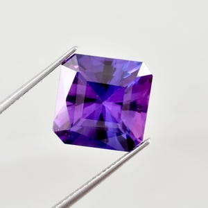 Puede incluir: Una gema púrpura facetada de corte cuadrado sostenida por unas pinzas. La gema tiene un color profundo y rico con reflejos internos. Las facetas crean un juego de luces y sombras, realzando su brillo. La gema está sobre un fondo blanco.