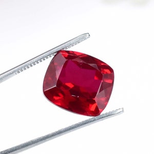 May include: A deep red gemstone with a cushion cut, held by tweezers against a white background.