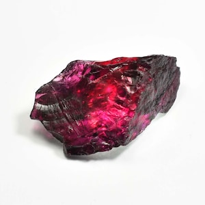 May include: A rough-cut, natural gemstone with a deep red and purple hue. The raw mineral has a crystalline structure and a textured surface, reflecting light. The gemstone is set against a white background.