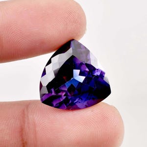 Puede incluir: Una gran piedra preciosa de amatista triangular facetada con un tono púrpura profundo. La piedra preciosa está tallada en forma triangular con muchas facetas que reflejan la luz.