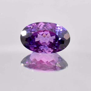 以下が含まれることがあります： 深い紫色のオーバルカットの宝石。光と影のコントラストが美しい。宝石のファセットは輝きを生み出し、紫とラベンダーの色合いを反射しています。反射する表面に置かれています。