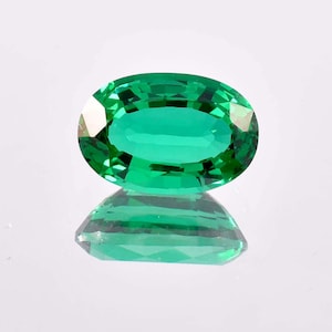 Puede incluir: Una gema de esmeralda tallada en forma ovalada y facetada. La gema es de color verde vibrante y refleja la luz, creando un efecto brillante. La piedra preciosa se muestra sobre una superficie reflectante, mostrando su imagen reflejada.