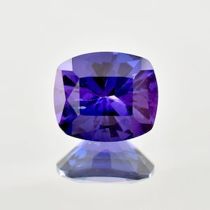 May include: A cushion-cut gemstone in a deep purple shade. The facets of the gem catch the light, producing a sparkling effect. The gem is set against a white backdrop, with its reflection visible beneath it.