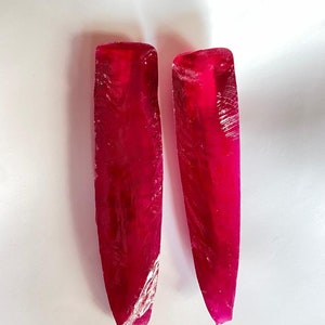 Puede incluir: Dos piedras preciosas de rubí largas, delgadas y facetadas, ambas de un color rojo intenso y rico. Las piedras preciosas están talladas en forma rectangular con extremos puntiagudos.