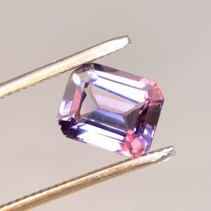 May include: A purple and blue gemstone with a rectangular emerald cut. The gemstone is held in tweezers.