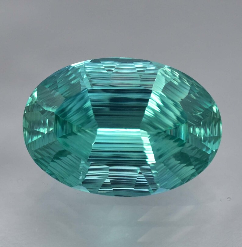 May include: A vibrant teal blue gemstone with a faceted oval cut. The gemstone has a striated pattern and a high polish.