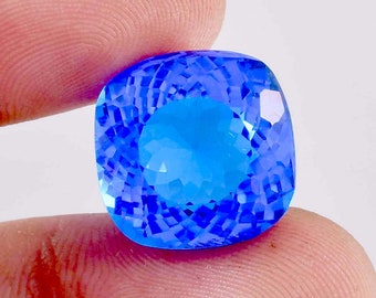 Può includere: Una grande gemma sfaccettata con taglio a cuscino quadrato. La gemma è di un colore blu vibrante, con tonalità blu più chiare al centro. La gemma è tenuta tra due dita su uno sfondo bianco.