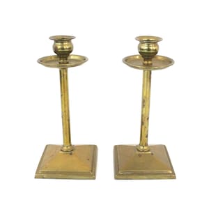 Può includere: Un paio di candelabri in metallo dorato con base quadrata e coppa rotonda per candele. I candelabri sono su uno sfondo bianco.