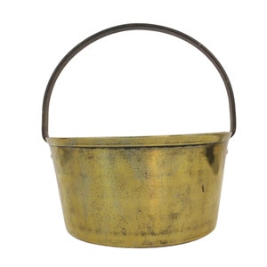 May include: An antique brass bucket with a dark metal handle. The bucket has a slightly tapered shape and a tarnished, aged appearance, suggesting it is vintage. The handle arches over the top, providing a way to carry the bucket.
