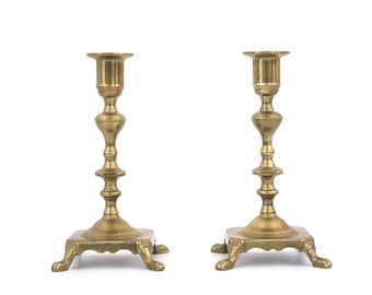 May include: A pair of gold metal candle holders with a classic design. Each holder has a square base with three legs and a tall, slender stem with a cup at the top.