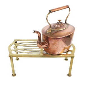 Puede incluir: Una tetera de cobre con asa de latón se asienta sobre un soporte de latón con patas decorativas.