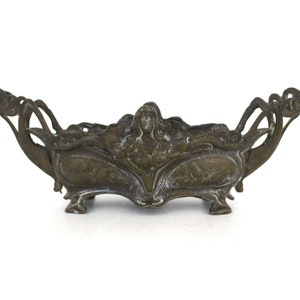 May include: An ornate brass planter with a decorative design featuring a woman's face and floral motifs. The planter has four legs and is designed to hold a small plant.