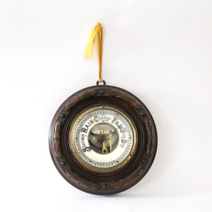 May include: A vintage wooden framed barometer with a white face and gold accents. The face has the words "Storm Rain" and "Fair Very Dry" printed on it. The barometer is hanging from a yellow ribbon.