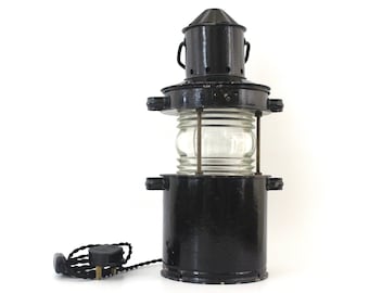 Op de afbeelding: Een zwarte metalen vuurtorenlamp met een heldere glazen lens en een zwart elektrisch snoer.